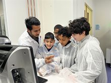 Four young students wearing lab coats watching intently to a researcher holding a chip package