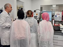 Three young girls seen from the back listening to a scientist who is explaining a wirebonder