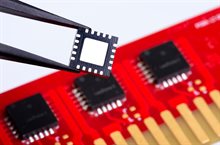 Chip between tweezers in front of a red PCB with other chips on it